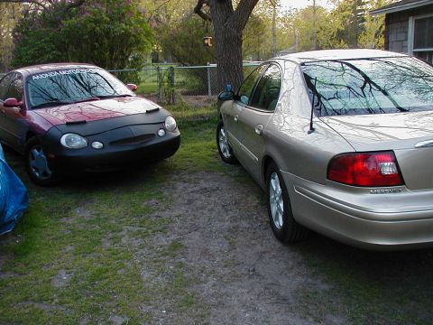 1997 Ford Taurus - A red 1997 Ford Taurus and a tan Mercury Sable are parked side-by-side on a gravel driveway.