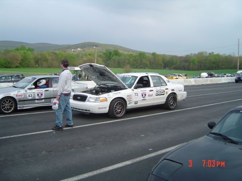 1999 Ford Crown Victoria - Two race cars are lined up on a track, one with its hood open, under a cloudy sky.