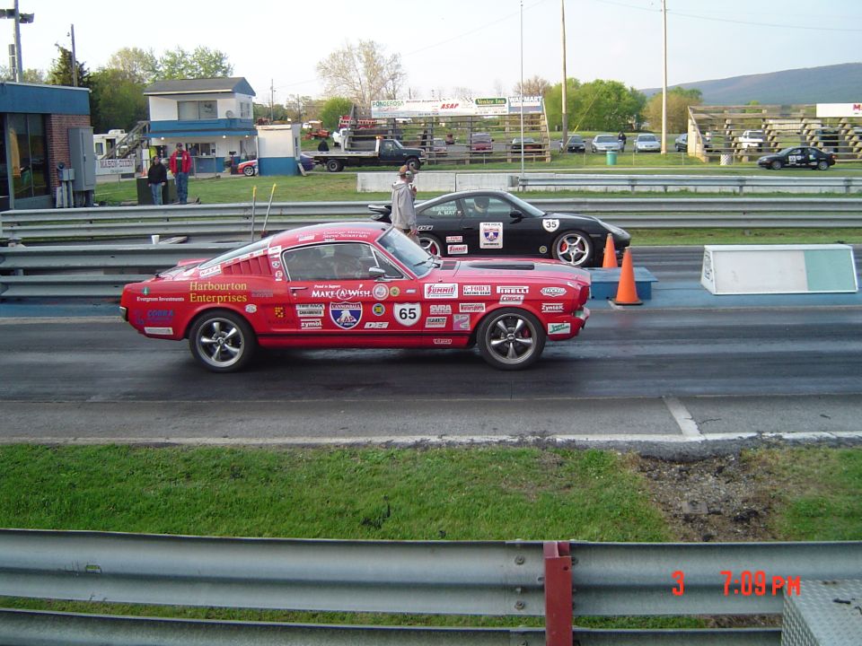 Red 1960s Ford Mustang race car with numerous sponsor decals and number 65, alongside a black Porsche race car with number 35, on a drag strip