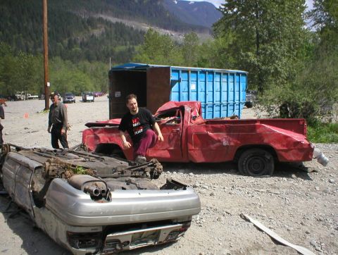 1978 Ford Bronco - Two heavily damaged vehicles are in a junkyard setting with a large blue dumpster nearby.