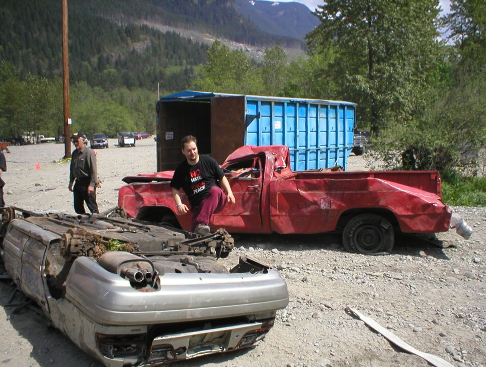 Guy in a t-shirt sits on a wrecked red pickup next to an overturned car. Blue dumpster in background