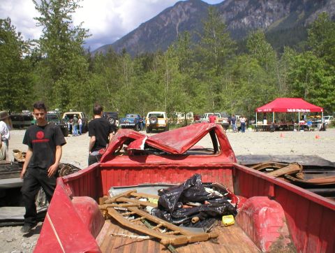 1978 Ford Bronco - The rear of a red pickup truck is filled with trash and debris, with a gathering of people and vehicles in the background.