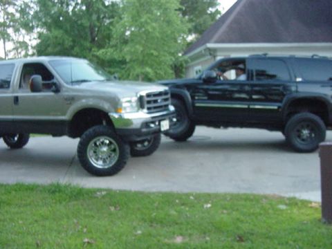 2000 Chevrolet Tahoe - Two lifted SUVs, one black and one silver, are parked side-by-side on a paved surface in front of a house.