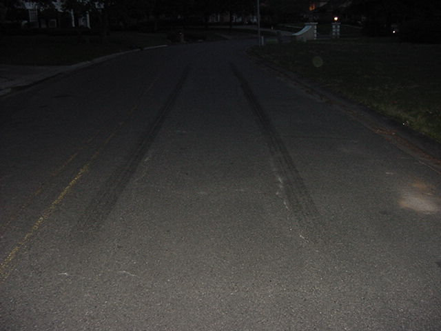 Tire tracks on a dark asphalt road at night, with a faint yellow line visible on the left