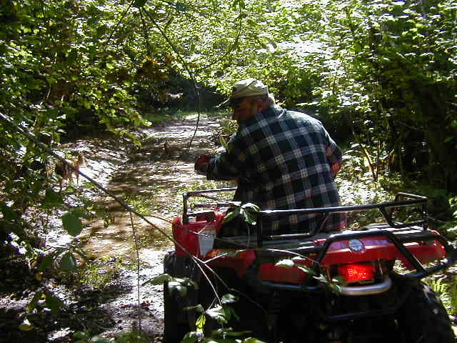 Red ATV on muddy forest trail with man in plaid shirt standing beside it