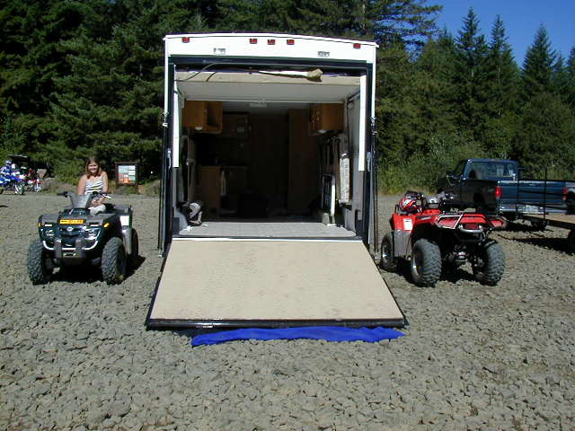 White cargo trailer with open rear ramp between a green and red ATV on gravel