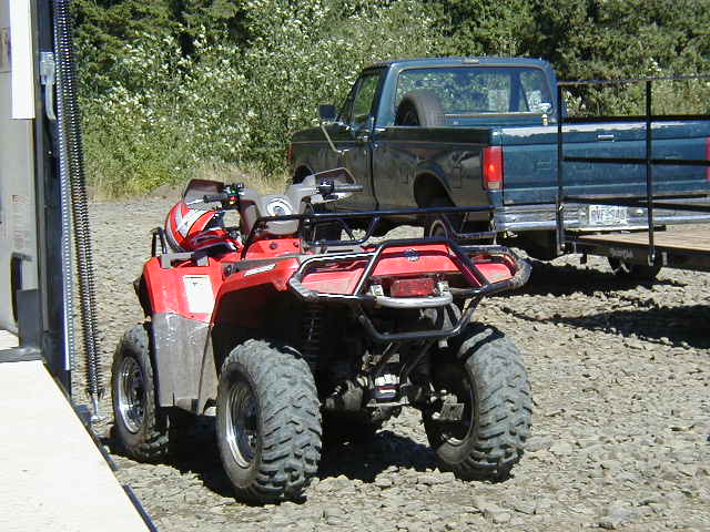 Red ATV parked on gravel with helmet on front rack, teal pickup and trailer behind