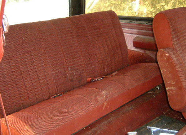 Red fabric bench seat and bucket seat interior of a dusty vehicle, likely from the 1980s