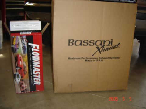 1990 Ford Bronco - Two boxed exhaust parts sitting on a garage floor: a red Flowmaster muffler carton opened at the top next to a large plain Bassani Xhaust b…