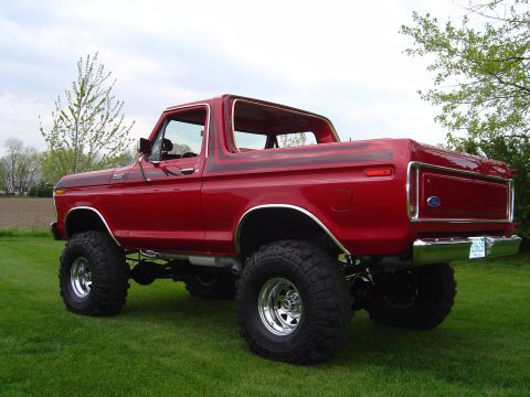 1978 Ford Bronco - Three-quarter rear view of a lifted candy-red full-size Ford Bronco with its hardtop removed, parked on a green lawn with a plowed field be…