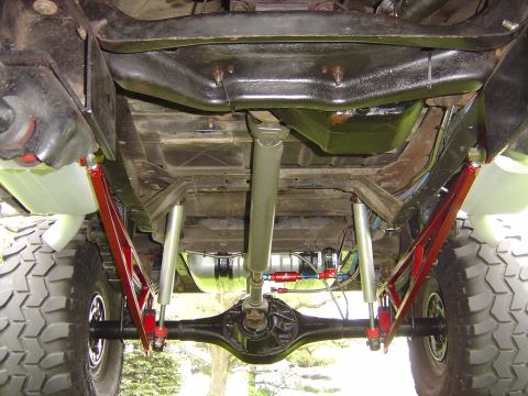 1978 Ford Bronco - Undercarriage view from behind a lifted 4x4, showing the black rear axle housing, driveshaft, red-painted four-link ladder bars and shocks,…