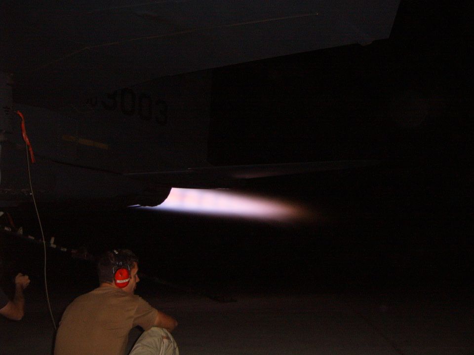 Jet engine afterburner ignites, emitting a bright pinkish-purple flame. A person wearing headphones watches from below