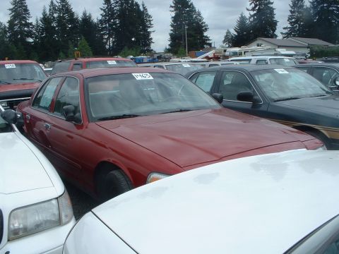 1999 Ford Crown Victoria - A red sedan is parked among other vehicles in an outdoor lot under a cloudy sky.