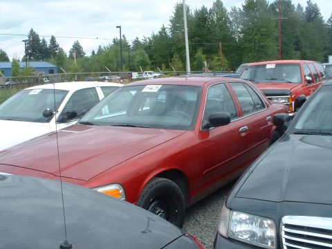 1999 Ford Crown Victoria - A red sedan is parked in a lot with other vehicles, including a white sedan and a red SUV visible in the background.