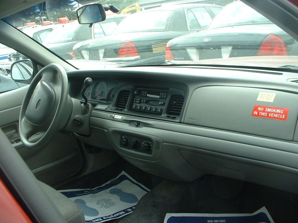 Interior view of a 1998 Ford Crown Victoria showing dashboard, steering wheel, and "No Smoking In This Vehicle" sticker. Other cars visible through the windshield