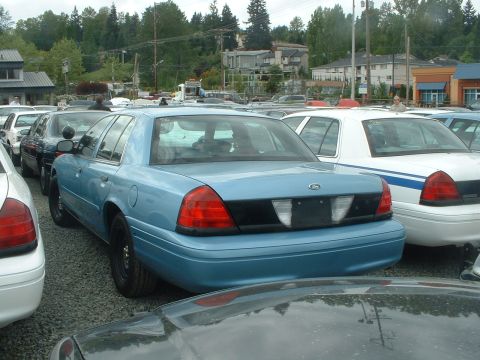 1999 Ford Crown Victoria - A blue sedan is parked in a lot with other vehicles, including a white car with blue stripes.