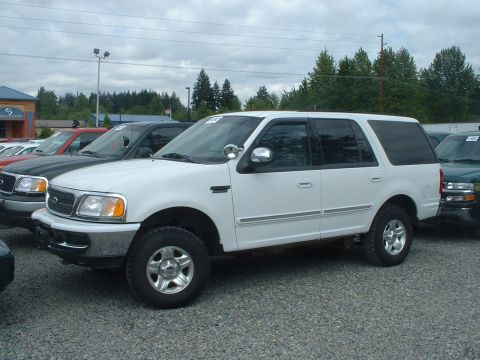 1999 Ford Crown Victoria - A white SUV is parked in a lot with other vehicles under a cloudy sky.