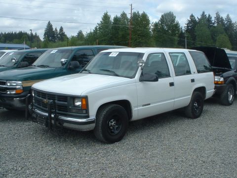 1999 Ford Crown Victoria - A white SUV with a push bar is parked in a gravel lot with other vehicles.