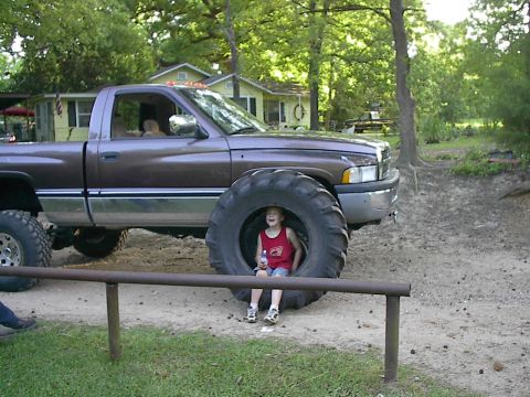 1997 Dodge 1500 - A child sits inside the massive front tire of a lifted pickup truck, with houses and trees in the background.