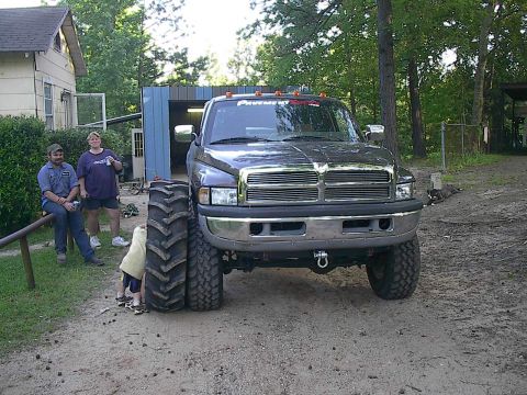 1997 Dodge 1500 - A dark gray 1997 Dodge 1500 pickup truck is parked outside a garage with two people watching a child near its massive tires.