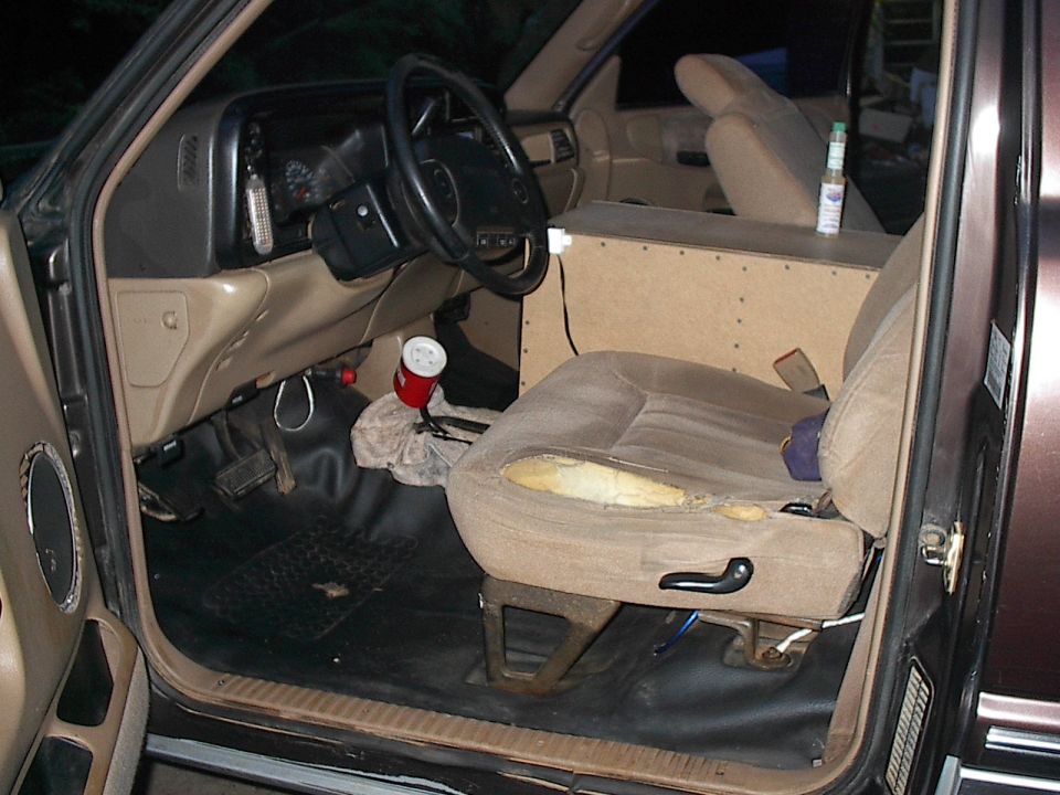 Pickup interior through open driver's door showing tan dash, torn seat cushion, and custom wood box between seats