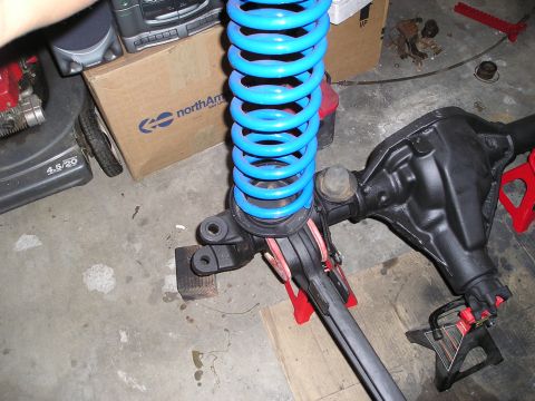 1986 Ford Bronco II - A front axle assembly with a bright blue coil spring is shown, supported by jack stands in a garage setting.