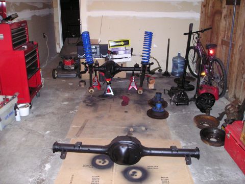 1986 Ford Bronco II - Various vehicle axles and suspension components are organized on a garage floor, suggesting a project in progress.