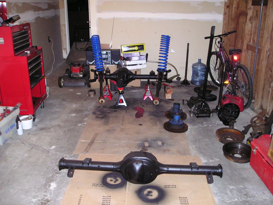 Vehicle axles and suspension parts laid out on a garage floor with a red toolbox and lawnmower in the background