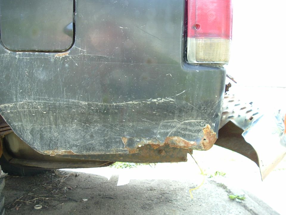 Dark-colored vehicle's rusted rear bumper and lower quarter panel with visible scratches and damage