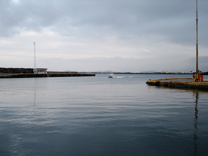 White motorboat entering a calm harbor between breakwaters under overcast sky.