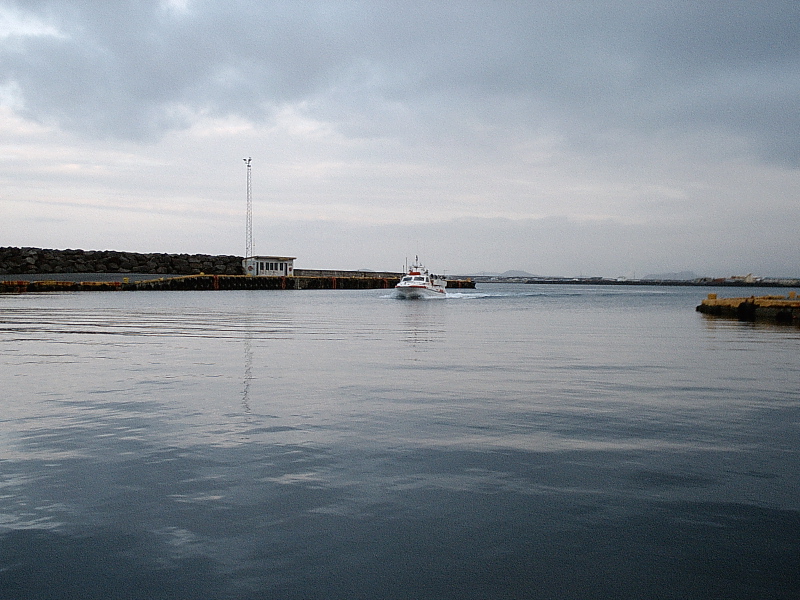 Small white boat entering a calm harbor past a breakwater on an overcast day