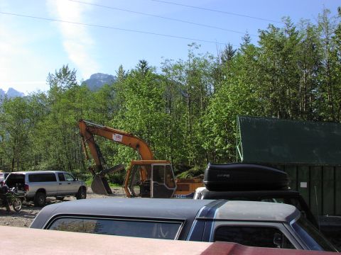1993 Ford Bronco - An excavator sits idle in a lot with several vehicles, surrounded by trees and a mountainous backdrop.