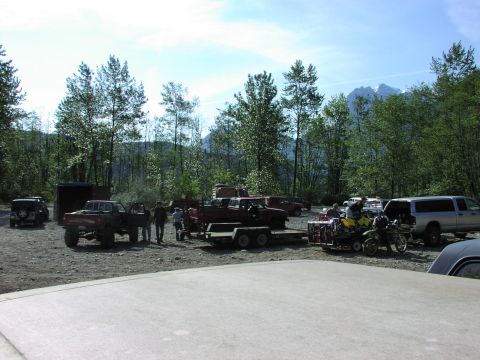 1993 Ford Bronco - A collection of vehicles and people are gathered in a wooded area, with mountains visible in the distance.