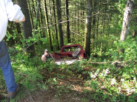 1993 Ford Bronco - A red pickup truck from the 1980s is seen abandoned in a wooded area, with a man visible in its bed.