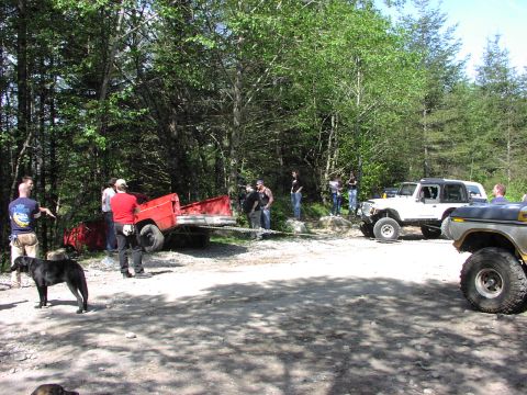 1993 Ford Bronco - Several off-road vehicles and people are gathered in a wooded area, with a red trailer being moved.