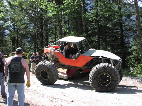 1993 Ford Bronco - A custom-built off-road buggy with oversized tires and a prominent roll cage is seen on a dirt path, with other vehicles and people in the…