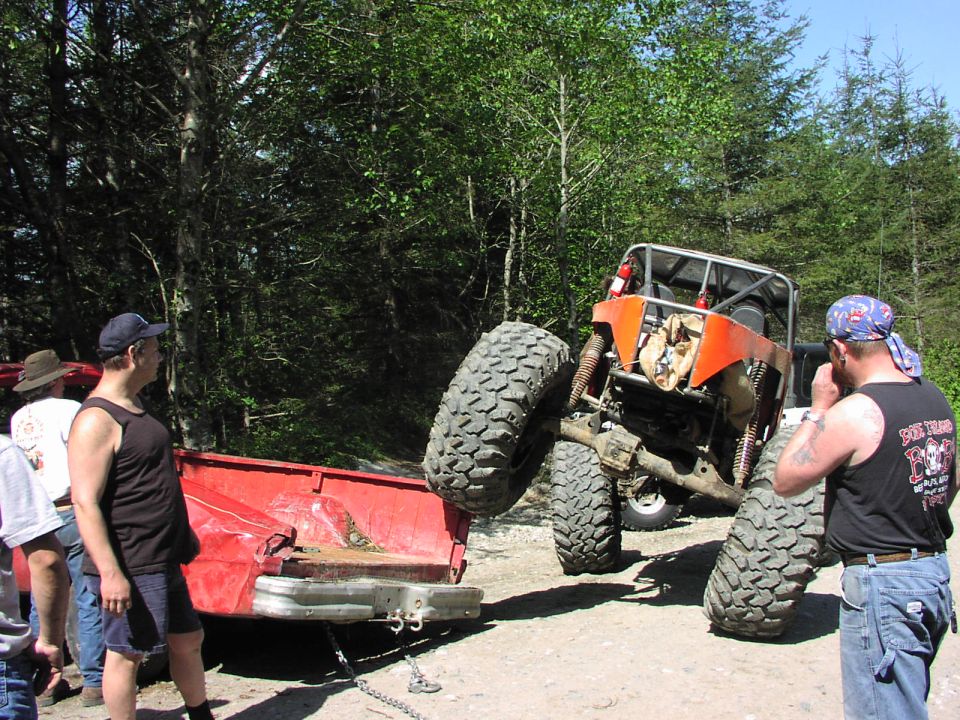 Orange off-road buggy with large tires is stuck, being pulled by a red pickup truck. People watch