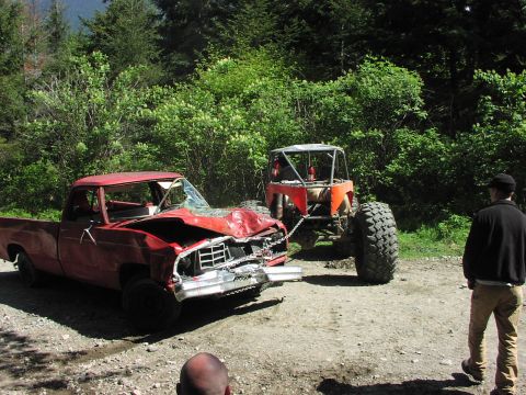 1993 Ford Bronco - A severely damaged red pickup truck is being towed by a large off-road vehicle on a dirt road.