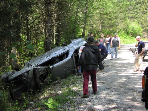1993 Ford Bronco - A group of people gather around a silver SUV that has crashed and overturned in a wooded area.