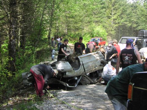 1993 Ford Bronco - A group of people work to recover a heavily damaged SUV from a wooded area, with other vehicles present.