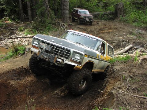 1993 Ford Bronco - Two Ford Broncos off-roading on a dirt path with trees and logs surrounding them.