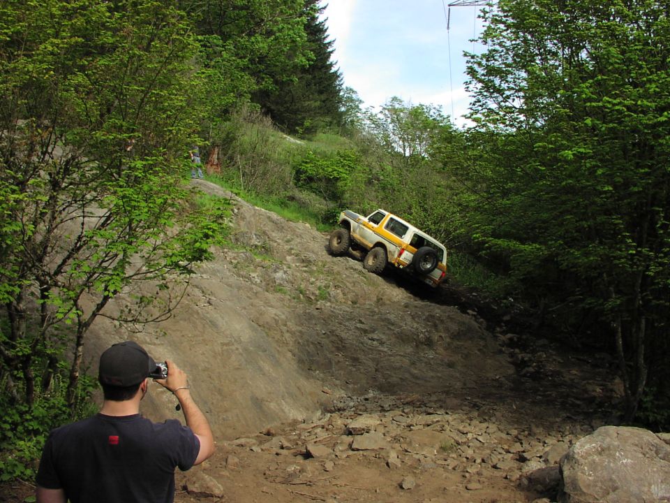White and yellow Ford Bronco climbing a rocky incline, with a person photographing it from below