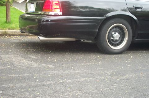 1999 Ford Crown Victoria - Close-up of the rear driver-side quarter of a black Ford Crown Victoria on a street, showing a stainless exhaust tip exiting beneath the re…