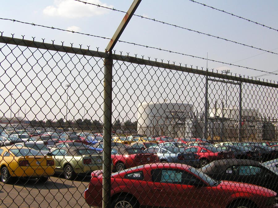 Red 2005 Ford Mustang parked in a lot behind a chain-link fence with barbed wire