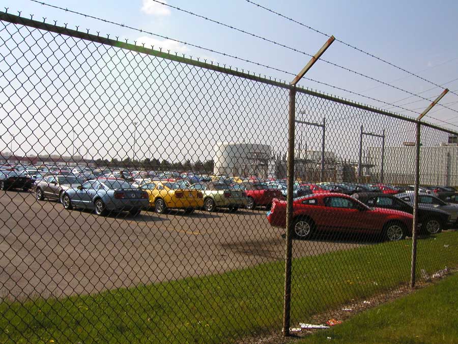 Red 2005 Ford Mustang parked in a lot with many other cars, viewed through a chain-link fence