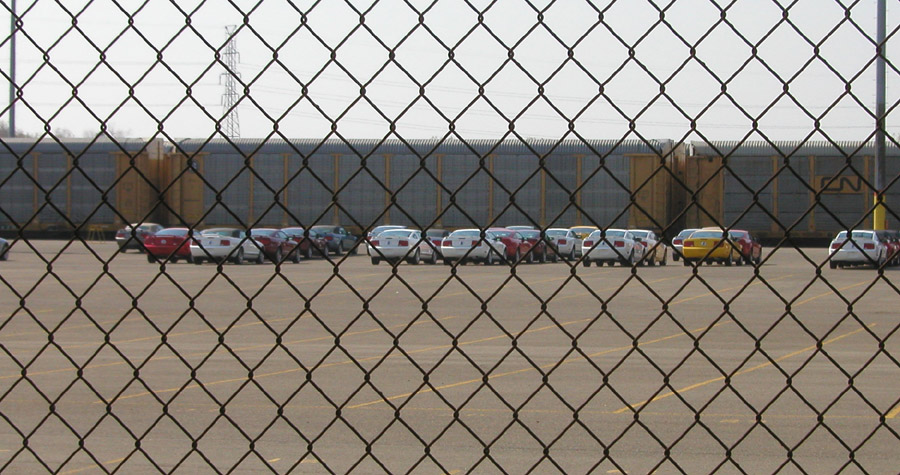 Cars parked in a lot with a train in the background, viewed through a chain-link fence