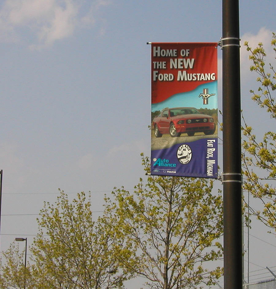 Banner on pole says 'Home of the New Ford Mustang' with a red car. Also shows 'Auto Alliance' and 'Flat Rock, Michigan'