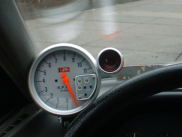 Dash-mounted white-face 11,000 RPM tachometer with orange needle above steering wheel