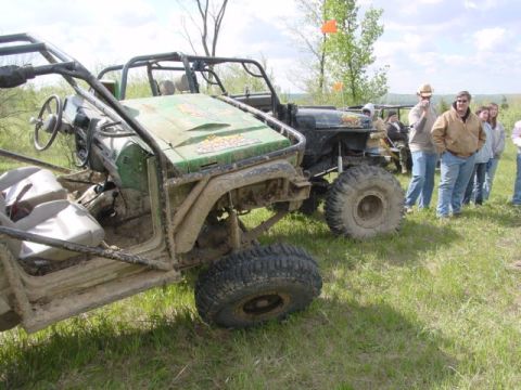 1992 Ford Bronco - Two heavily modified off-road vehicles sit in a grassy field, covered in mud.