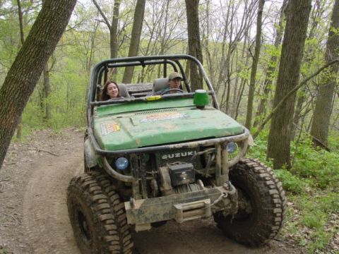1992 Ford Bronco - Two people ride in a modified green Suzuki off-roading on a dirt path through a wooded area.